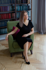 Blondie long hair woman is sitting on a cozy olive chair and drinking coffee.