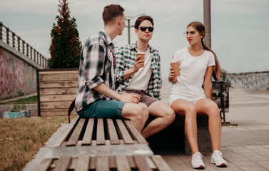 Fototapeta premium The Company Of Young Guys Rest And Drink From Paper Glasses Sitting On A Bench During A Summer Journey.