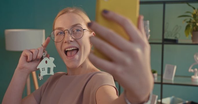 Close up of Caucasian blonde happy pretty young woman showing off keys from new apartment to phone camera and taking selfie photo. Female real estate owner making selfies photos in cozy living room.