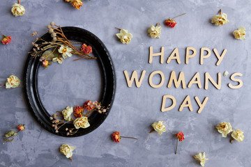 Dried flowers, sprigs, leaves are scattered inside black vintage oval frame and around on concrete cement background. Wooden inscription happy women day. Greeting card template, invitation.
