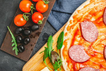 Process of making pizza round dough with black olives, cherry tomatoes, mozzarella cheese, sliced salami and green leaves spinach, rucola, top view