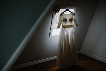 The bride's dress hangs on the wall by the window.