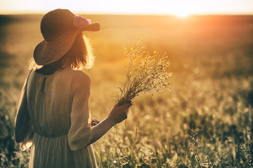 Stylish woman with hat holding grass herb bouquet in summer field enjoying morning sunrise or...