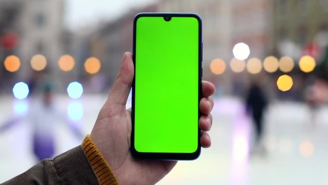 Green screen smartphone on ice rink with city lights on blur background. Chroma key mockup display for free content. Online connection. Stream trend. Winter holidays abroad. Happy feelings.