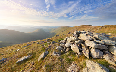The summit cairn of Rampsgill head at sunset with views of Ramps Gill and Martindale Common in the evening sun in the Lake District.