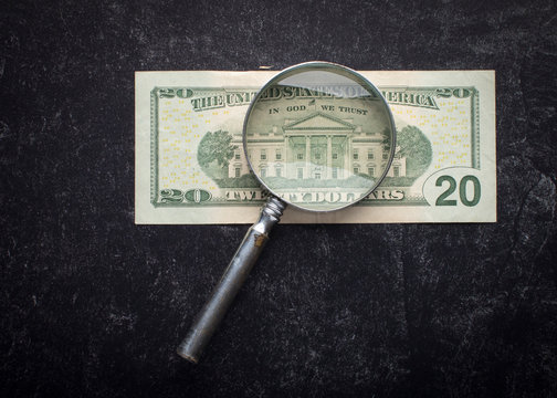 Antique magnifying glass highlights the white house and the words In God We Trust on the back of the twenty dollar bill on a black slate background