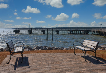 Fototapeta na wymiar Two empty benches on a patio by a pier over sun dappled sea