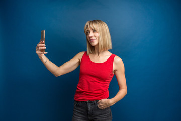 Fototapeta premium Beauty fashion portrait of slim blonde woman in red shirt on a blue background in studio making a selfie with smartphone. Social networks, selfie addiction