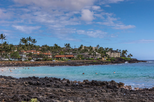 Poipu, Kauai, Hawaii, USA. - January 11, 2012: Black rocks form Brenneckes beash and bay with blue ocean water in front of Poipu shores with large mansions, green trees, under blue cloudscape.
