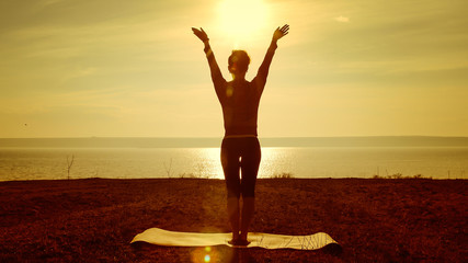 Young woman silhouette practicing yoga on the sea beach at sunset. Slim woman doing gymnastics over morning sun. Girl with a slender figure practices yoga near the ocean at sunrise. Sport and health.