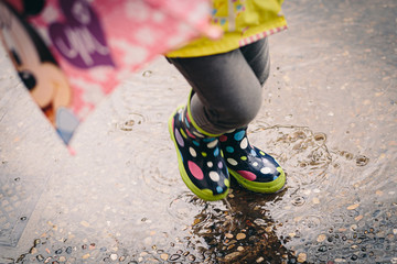 Colorful spotted rain boots baby girl playing with water puddles on a urban scene