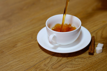 Classic white cup of black tea with saucer, teapot, sugar and cinnamon on the wooden table background. Pouring tea. Breakfast drink at home or cafe.