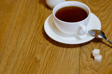 Classic white cup of black tea with saucer, teapot, sugar and cinnamon on the wooden table background. Breakfast drink at home or cafe.