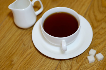 Classic white cup of black tea with saucer, teapot, sugar and cinnamon on the wooden table background. Breakfast drink at home or cafe.