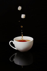 White cup with black tea on black background. Sugar falling in cup of hot aromatic black tea with splashes . Energy drink for breakfast.