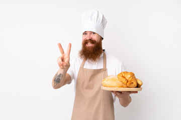 Redhead man in chef uniform. Male baker holding a table with several breads smiling and showing victory sign