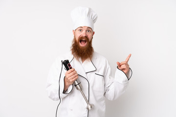 Redhead man using hand blender over isolated white background pointing finger to the side