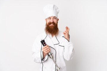 Redhead man using hand blender over isolated white background with fingers crossing