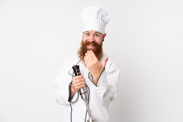 Redhead man using hand blender over isolated white background inviting to come