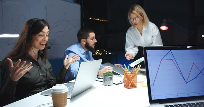 Pretty young Caucasian woman working at computer in call center and talking with client via web cam. Female worker of call-center late in evening. Co-workers speaking and having discussion behind.