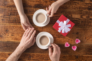 Valentine's Day celebration concept. A nice gift for your loved one. Hands of man and woman with coffee mugs on a wooden table background. Copy space. Flat lay. Close-up.