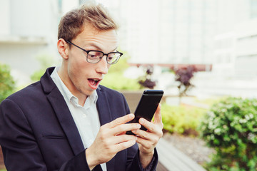 Excited office employee with cellphone getting shocking news. Amazed business man in eyeglasses and jacket staring at phone screen with wide eyes and open mouth. Unexpected news concept