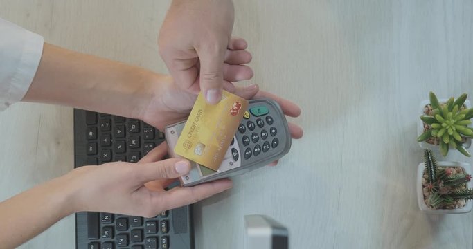 Bird's Eye View Shot Of Hands Over Counter Making A Contactless Payment With Gold Credit Card And POS Terminal.