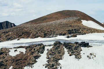 Beautiful small glacier with ice cornice on stony hill under cloudy sky. Snow on mountain. Firn on...