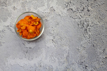 Dried carrot chips in a glass bowl on a gray concrete background. Organic natural food. Top view, flat lay.