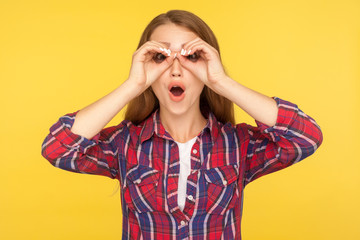 Portrait of surprised curious ginger girl in checkered shirt looking through hands on eyes, making binoculars gesture, expressing amazement and shock. indoor studio shot isolated on yellow background