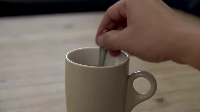 Hand pouring a tablespoon of sugar into a mug of coffee on a wooden table. Stirring a beverage with a spoon.