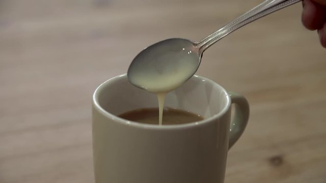 Hand pouring a tablespoon of condensed milk into a mug of coffee on a wooden table. Stirring a beverage with a spoon.