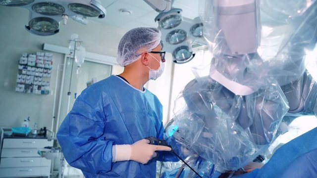 Medical specialist near the robotic device. Surgeon in blue protective uniform controls the work of medical robot on the screen.