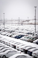 New cars covered with snow and parked in a distribution center on a cloudy day in the winter. A...