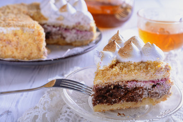 Piece of cake on a plate in the foreground and whole cake beautifully decorated with whipped protein peaks on a background next to a glass teapot and a cup of tea are ready for tea drinking