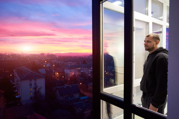 Side view of a man standing on a balcony with panoramic windows and looking into the distance while...