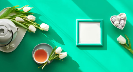 Spring panoramic flat lay. Tea cup, pot, sweets and white tulips on green. Valentine's day, Mother's day, international women day March 8. Diagonal sunlight, long shadows, trendy top view