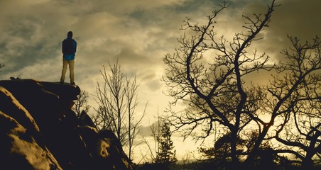 Male hiker with backpack on a mountain range behind a city looking at rugged winter landscape enjoying weekend. Old trees, cliff, sunset. Composite photo.  .