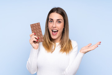 Young blonde woman over isolated blue background taking a chocolate tablet and surprised