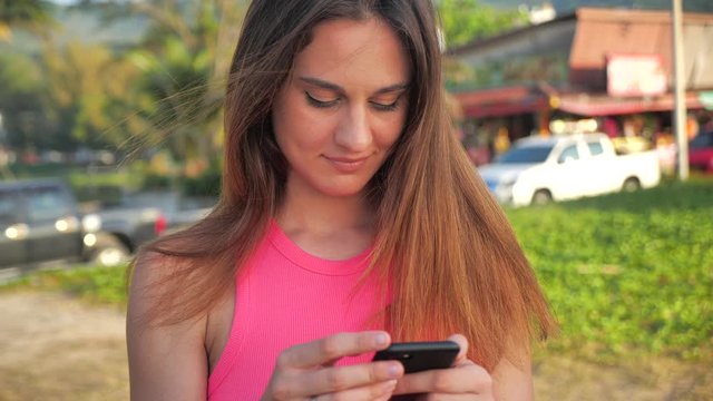 Close up portrait of an attractive girl in a pink short shirt. She stands in the site palms in the background. Cute is convicted, holding a smartphone in his hands. 4k
