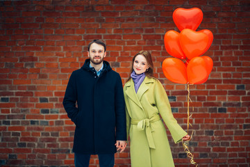 Obraz na płótnie Canvas happy caucasian lady and guy celebrating st valentines day together outdoors. couple dressed in coats enjoy spending time together, hold red air balloons. red brickwall in background. love concept