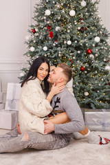 young beautiful couple, brunette girl in a white sweater, a blond man in a gray sweater sitting near a Christmas tree