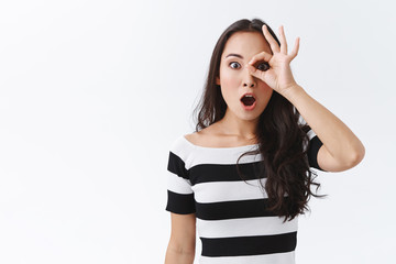Amazed good-looking brunette woman in striped t-shirt, gasping ambushed and amazed, look wondered through okay sign, making surprised expression, standing white background