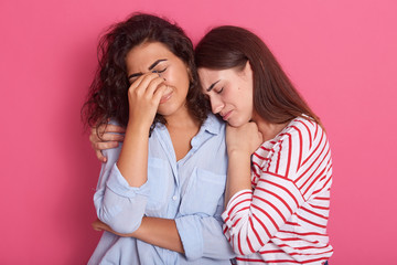 Close up portrait of one woman crying and another embracing her and calming down, female wearing blue shirt having problems, cries, keeping fingers on eyes, posing isoalted over pink studio background