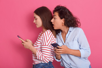Horizontal shot of two women sharing social media videos in smart phone indoors, attractive dark haired female looks at friend's phone screen with interest and widely opened mouth. Gossip concept.