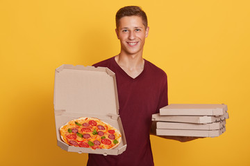 Horizontal shot of male holding picking tasty pizza, posing isolated over bright yellow studio background, attractive man wdresses casual maroon t shirt, looking at camera with charming smile.