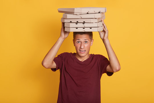 Indoor shot of courier with blonde hair, dresses casual burgundy t shirt holings many pizza boxes on his head, isolated over yellow studio background, looking directly at camera with astonishment.