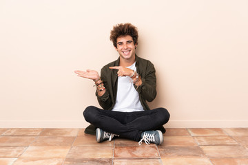 Young caucasian man sitting on the floor holding copyspace imaginary on the palm to insert an ad