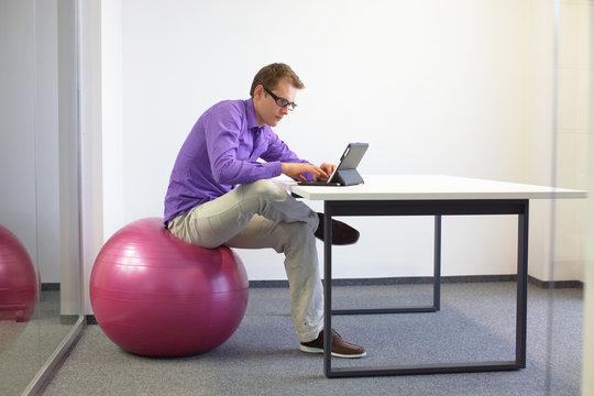 Man on stability ball working with tablet at desk in the office - changing siting  position