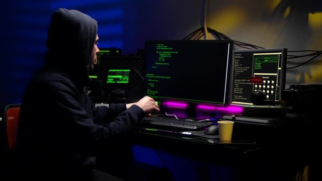 Male hacker working on a computer while green code characters reflect on his face in a dark office room.